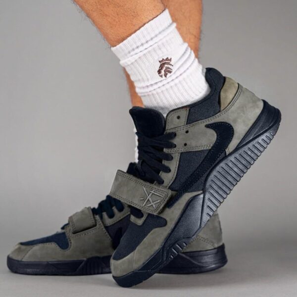 Close-up of a grey and black athletic sneaker worn with a white sock on a gray background, showing a chunky sole and layered panels.