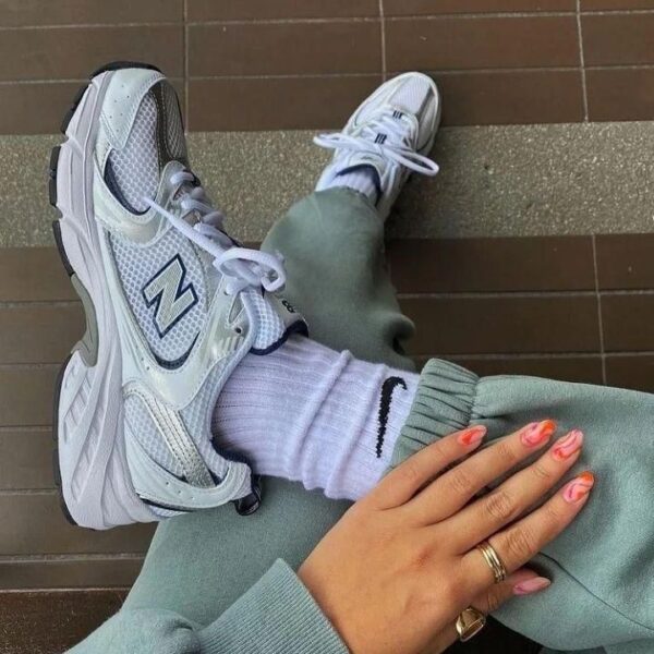 White New Balance sneakers with thick soles, white socks, sage-green joggers, and a hand with pink nail polish resting on the knee on a brown tile floor.