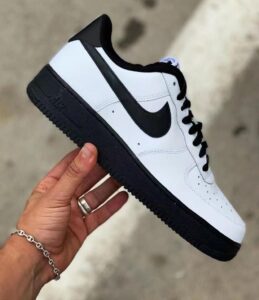 White leather Nike Air Force 1 sneaker with black swoosh and sole, held in a hand against a blurred outdoor background.