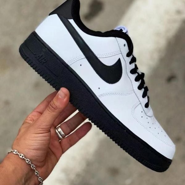 White leather Nike Air Force 1 sneaker with black swoosh and sole, held in a hand against a blurred outdoor background.