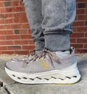 Close-up of gray On running shoes with yellow logo, worn over cuffed jeans, against a brick wall backdrop.