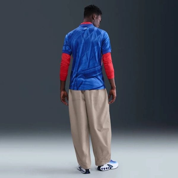 Back view of a person wearing a blue geometric jersey over red arm sleeves, beige wide pants, and slide sandals on a gradient backdrop.