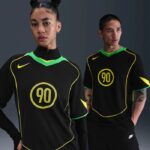 Two models wearing black athletic jerseys with neon yellow trim and a circular 90 emblem on the chest; athletic studio backdrop exists in gray.