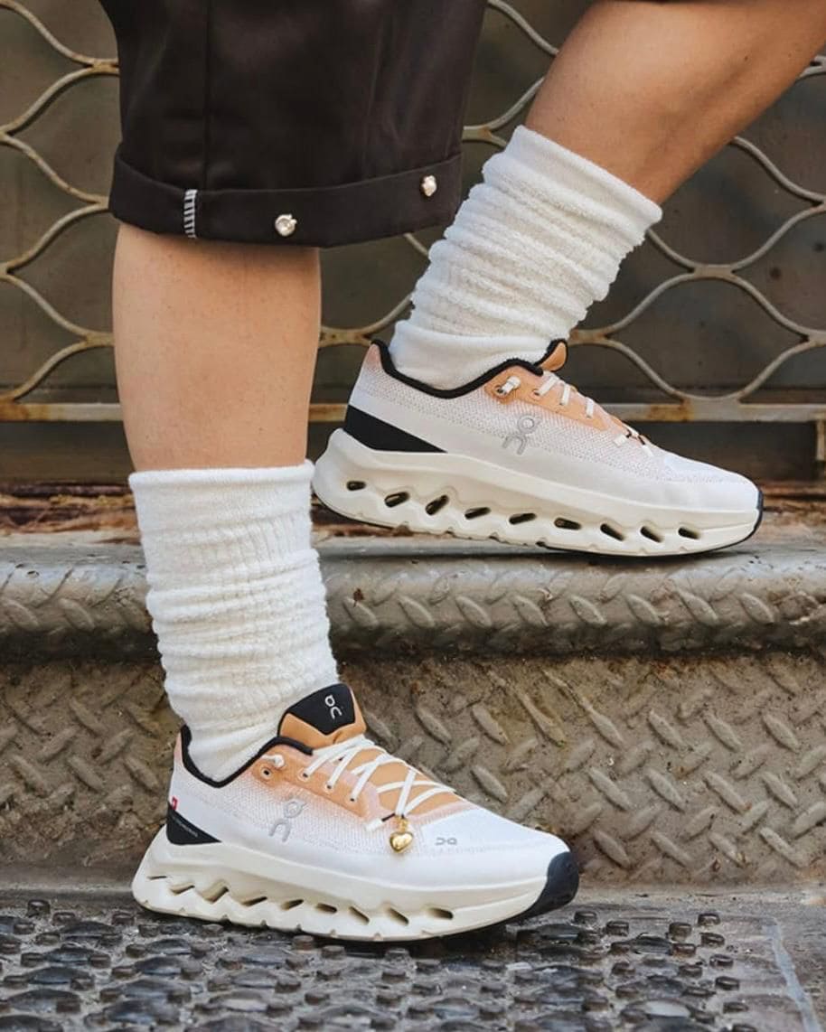 a80d6205-7d11-46c0-bad4-8f359b84b085 | UAE SPORT STORE Person wearing white and peach athletic sneakers with thick zigzag sole, white socks, standing on metal grate steps outdoors