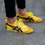 Pair of bright yellow sneakers with black stripes resting on a gray pavement surface, laces tied.
