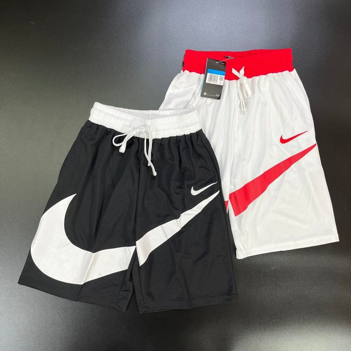 Two pairs of athletic shorts, black and white, with a red waistband and a tag attached on top of a dark surface.