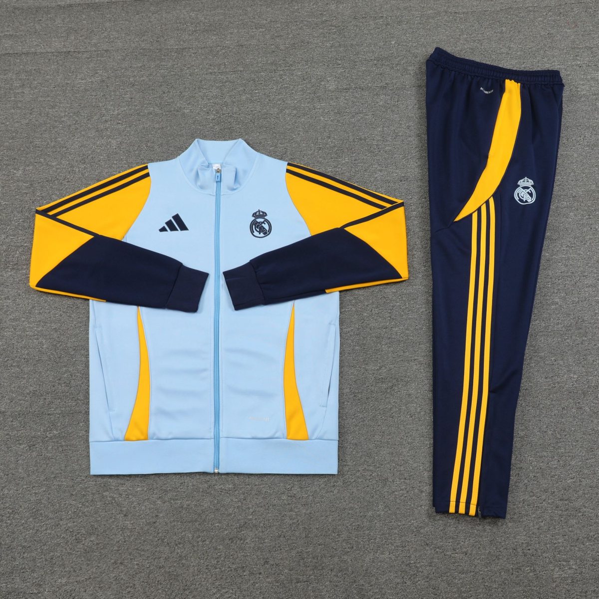 Light blue and navy Real Madrid tracksuit laid flat with yellow accents on a gray carpet. Crest and Adidas logo visible on the jacket sleeve patch and pants side stripes are yellow.",