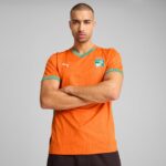 Man with arms crossed wearing an orange Ivory Coast national team soccer jersey, green trim, and a crest on the chest, against a gray studio backdrop.