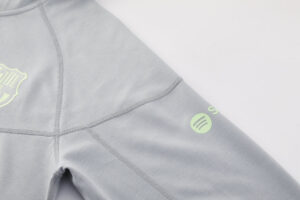 Close-up of a light gray sweatshirt sleeve with visible seam lines and a lime-green circular logo on the upper arm, plus a partial shield emblem on the chest area.