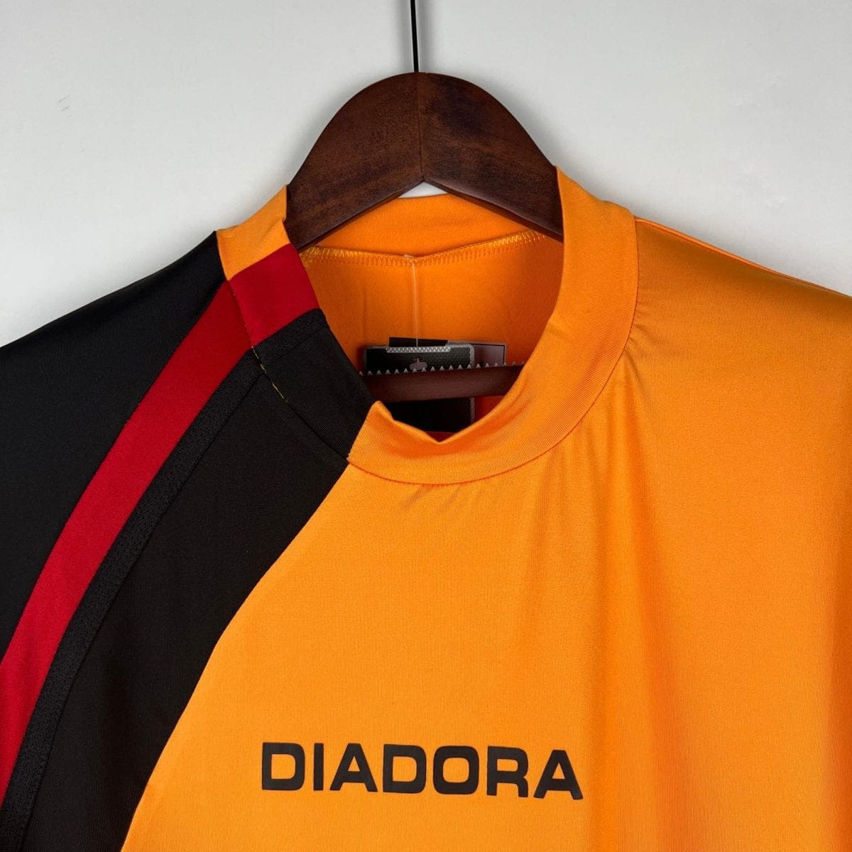 Orange sports jersey hanging on a wooden hanger, with black and red stripe details visible and the brand name DIADORA across the chest area.