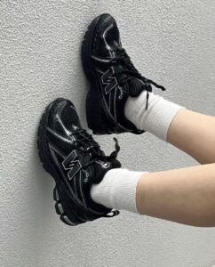 Two black athletic sneakers with chunky soles and white detailing rest against a textured gray wall, worn with white socks.