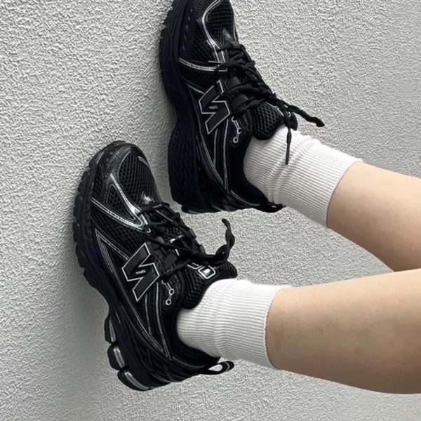 Two black athletic sneakers with chunky soles and white detailing rest against a textured gray wall, worn with white socks.
