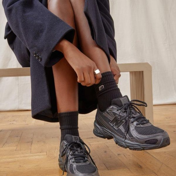 Person in a dark coat sits on a wooden floor and ties black athletic sneakers with chunky soles and black socks.