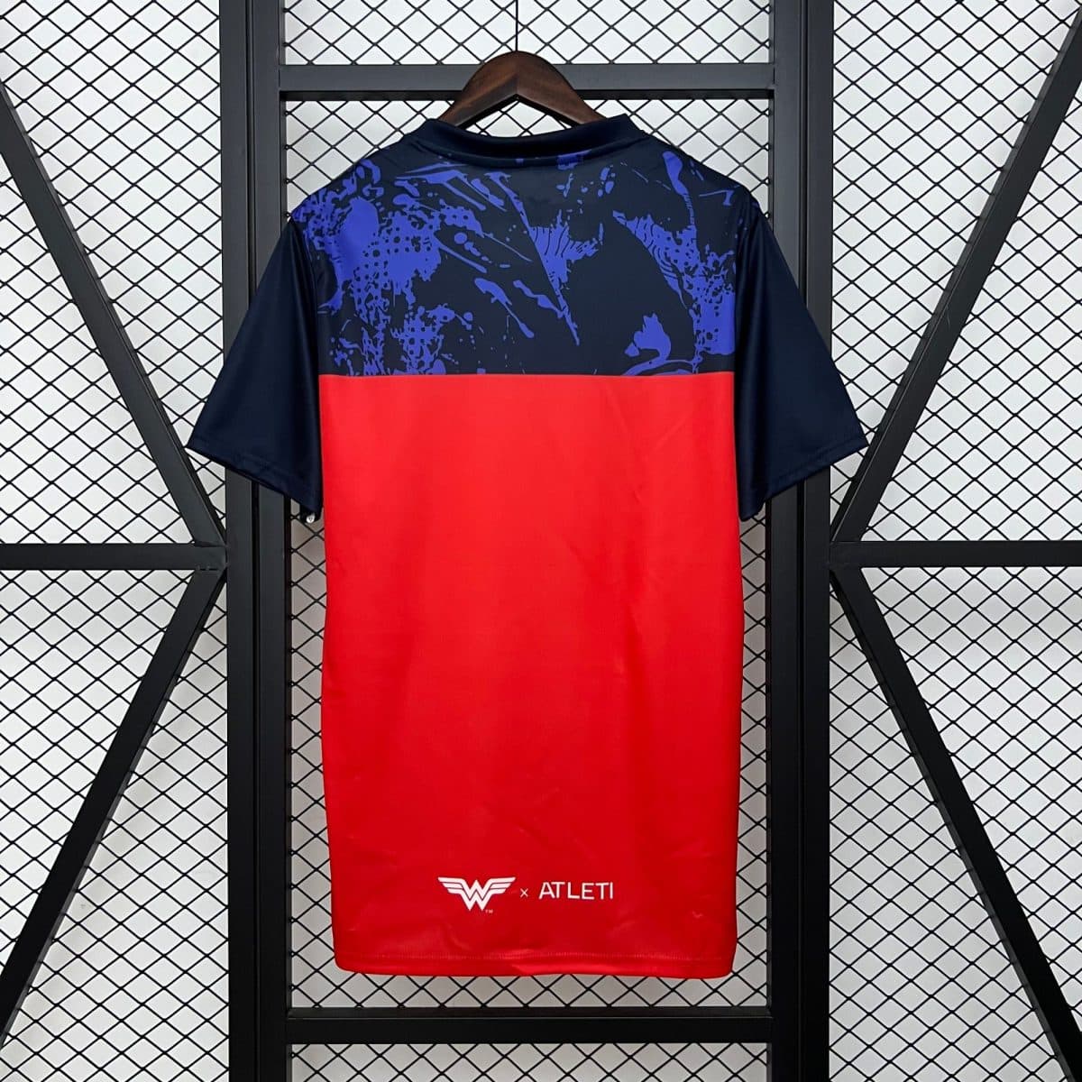 Red athletic jersey on a hanger with a navy-blue top featuring blue abstract splatter patterns and a bright red lower section; 'W x ATLETI' logo at the bottom hem.