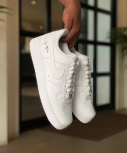Pair of white Nike Air Force 1 sneakers being held by a hand indoors, with a black-framed doorway in the background.