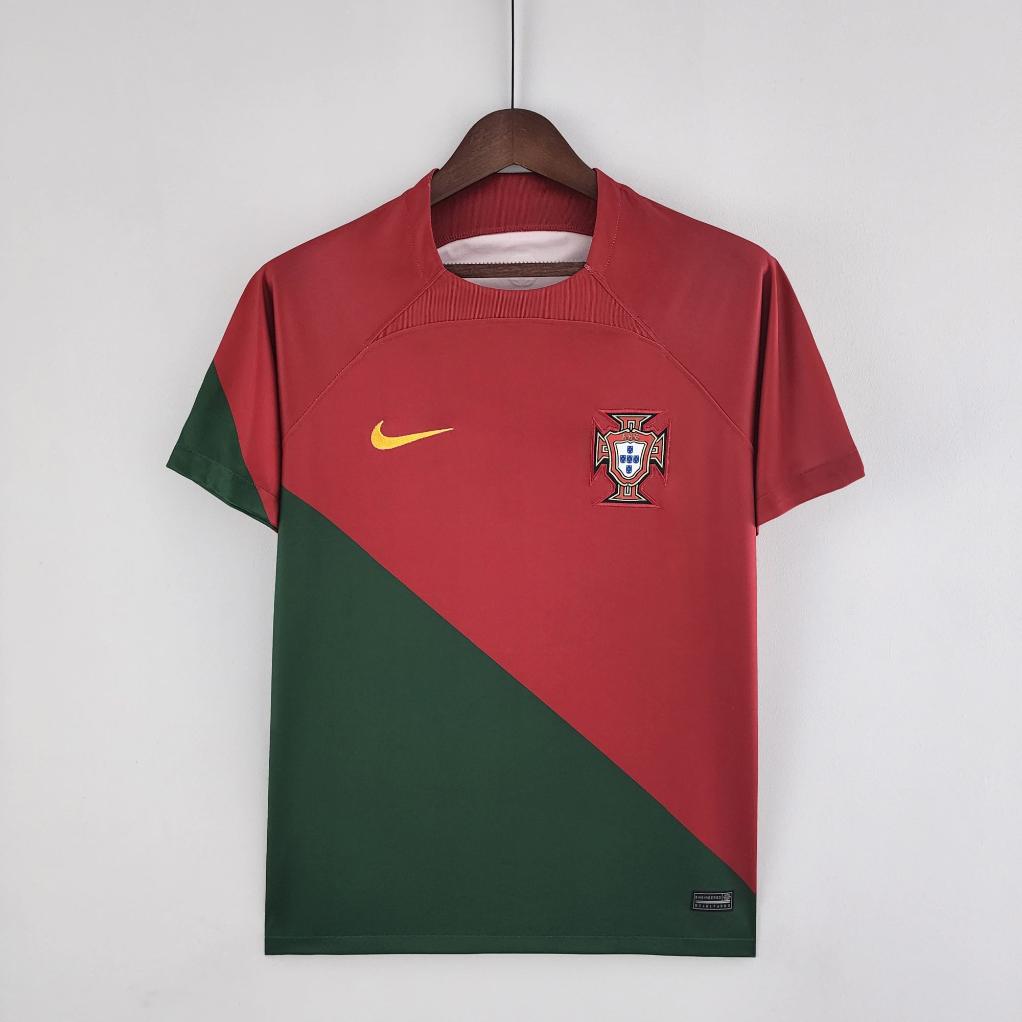 aa9619a0_b389442c-b324-4ccc-b1e9-9edfdf15c727 | UAE SPORT STORE Red Portugal national team soccer jersey on a wooden hanger, featuring a yellow Nike swoosh and crest on the chest, with a green diagonal panel on the side.