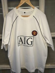 White Manchester United football jersey with AIG sponsor logo and club crest on the chest, hanging on a wooden hanger.