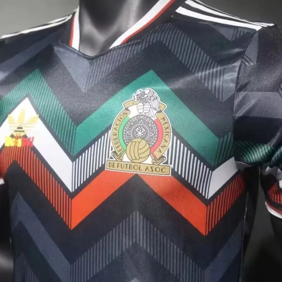 abb6195d3273793f | UAE SPORT STORE Close-up of a soccer jersey with a bold chevron pattern in black, green, red, and orange; center chest badge shows the Mexican Football Federation crest (Federación Mexicana de Fútbol).