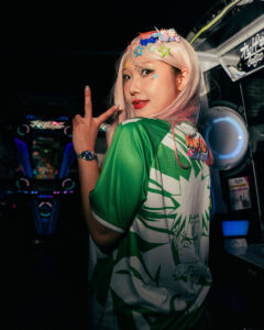 Woman with platinum blonde hair and star hair clips, wearing a green Naruto-themed jersey, posing with a peace sign in a dim arcade.