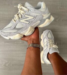Person holds a white and cream New Balance sneaker with beige laces, shown over a light wooden floor.