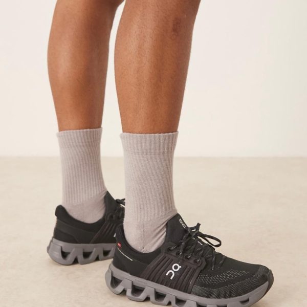 Lower legs with light gray crew socks in black ON running shoes on a pale floor, shown from the front side view.