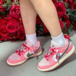 Kid wearing pink and white sneakers with rhinestone laces standing on concrete beside a wall of red roses