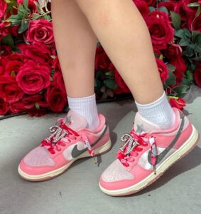 Kid wearing pink and white sneakers with rhinestone laces standing on concrete beside a wall of red roses