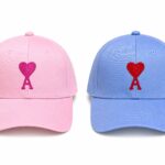 Two baseball caps side by side: pink cap on the left and light blue cap on the right, each with a small red heart-shaped ribbon embroidery on the front panel.