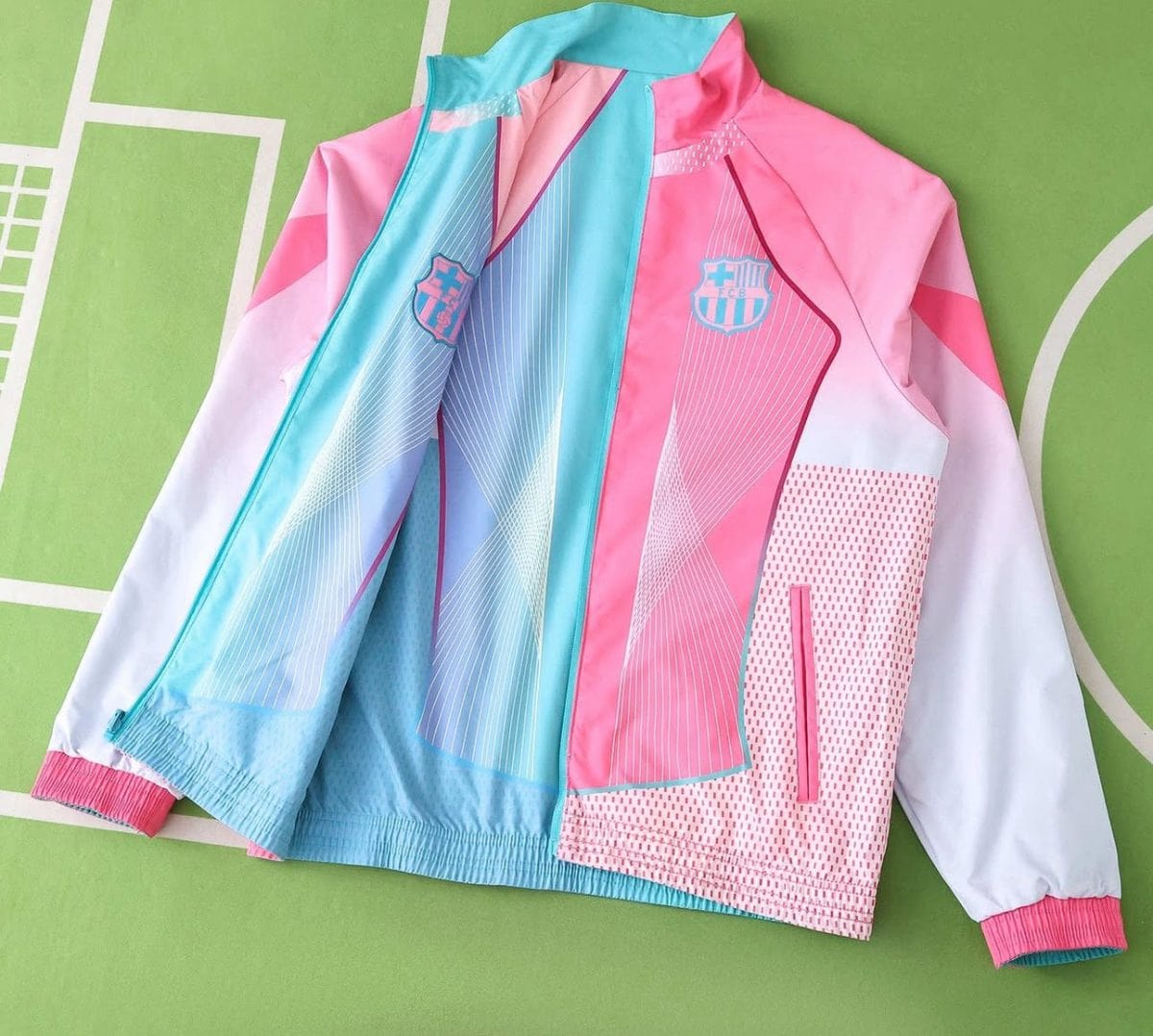 Pastel pink and blue sports jacket with FC Barcelona crest on the chest, white sleeves with pink cuffs, laid on a green background.