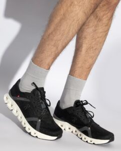 Black athletic sneakers with chunky white soles on a person wearing light gray socks, mid-step pose against a light background.