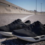 Pair of black athletic shoes with white soles resting on a sunlit asphalt surface beside a white lane line, railing and blue sky in background.