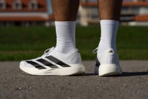 Close-up of white Adidas sneakers with black stripes and white socks standing on asphalt outdoors.