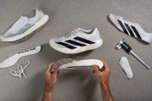 Hands holding a white sole while assembling a white athletic shoe on a gray work surface, with other shoe parts and tools nearby