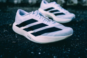 White Adidas Adizero running shoes with black stripes resting on a dark asphalt surface.