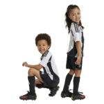 Two kids in matching black-and-white soccer uniforms posing against a white background; one crouching, one standing and smiling.
