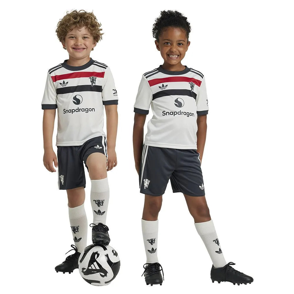 adidas-manchester-united-24-25-junior-third-set | UAE SPORT STORE Two smiling children in matching white soccer uniforms stand together with a soccer ball at their feet, ready to play.