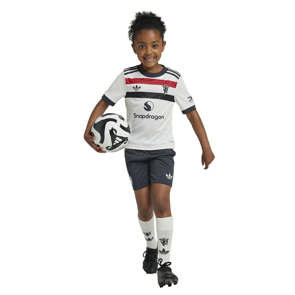 adidas-manchester-united-24-25-junior-third-set_4 | UAE SPORT STORE Young girl in a white soccer jersey with black and red stripes, dark shorts, and white knee-high socks holding a soccer ball and smiling.