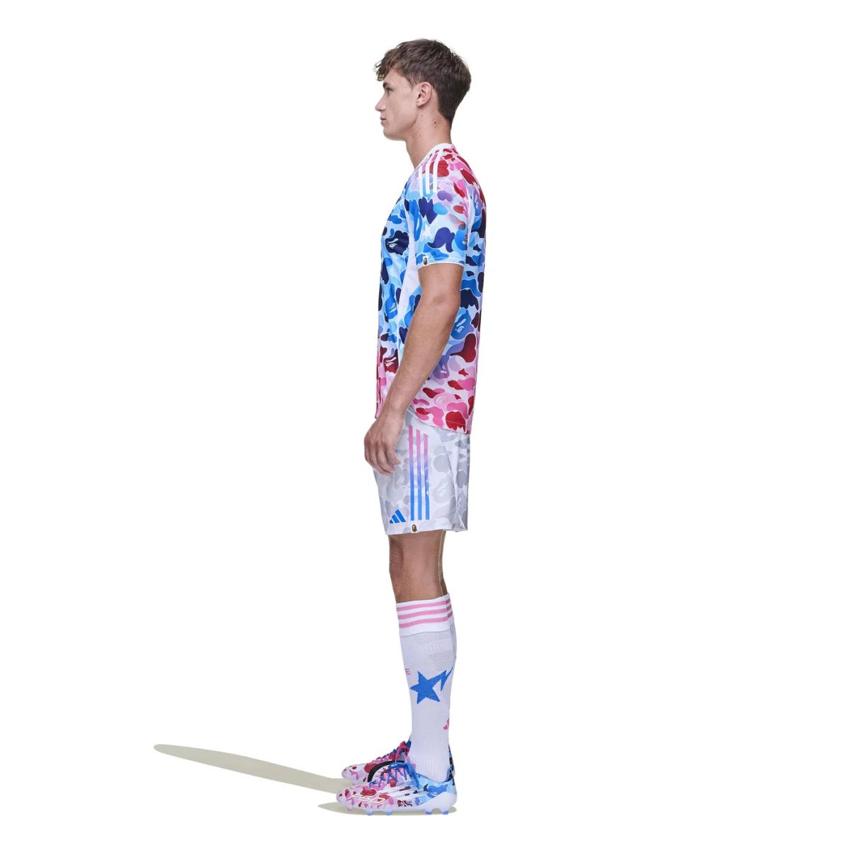 Side profile of a male soccer player modeling a pink-blue camouflage jersey, white shorts with blue stripes, and white socks with pink accents and stars.
