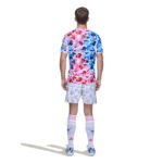 Back view of a person wearing a pink-to-blue camouflage soccer jersey, white camo shorts, white socks with pink stripes, and blue cleats, standing with shadow.