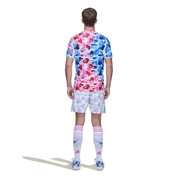 Back view of a person wearing a pink-to-blue camouflage soccer jersey, white camo shorts, white socks with pink stripes, and blue cleats, standing with shadow.