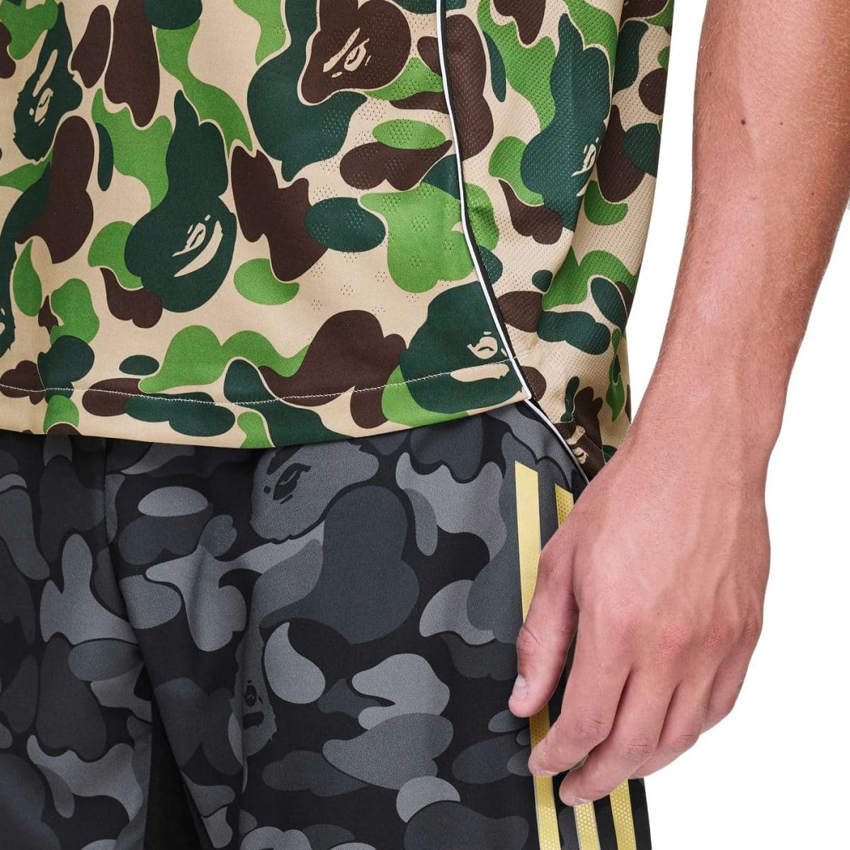 Close-up of a person wearing camo athleticwear: a green/brown camo jersey and gray camo shorts with a yellow side stripe, with the hand resting at the side.
