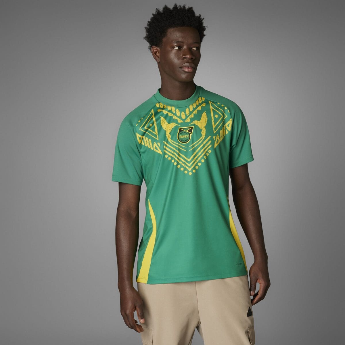 Male model wearing a green football jersey with a bold yellow geometric pattern and crest, standing against a gray backdrop.
