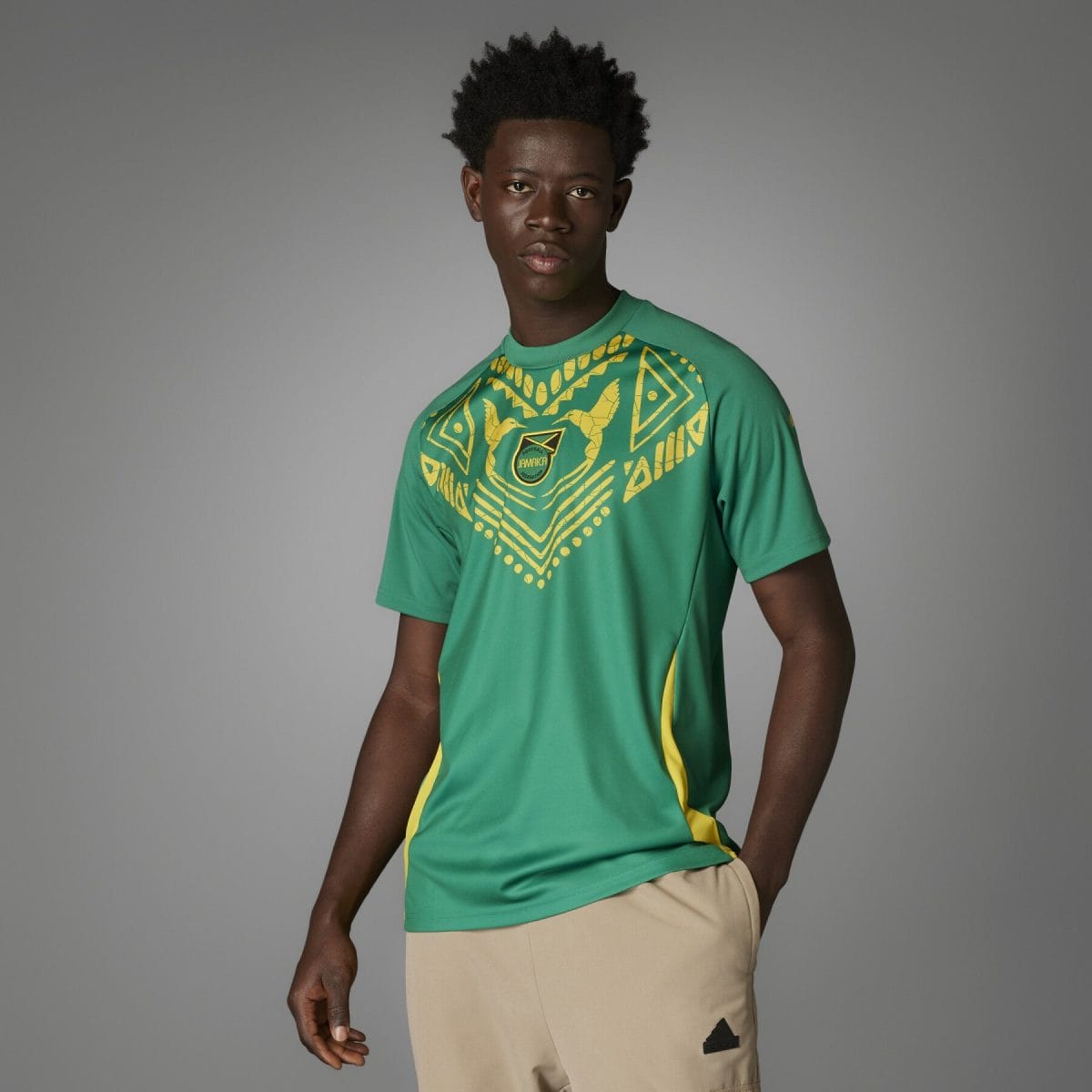 Young man wearing a green Jamaica football jersey with yellow geometric pattern, against a gray studio background.