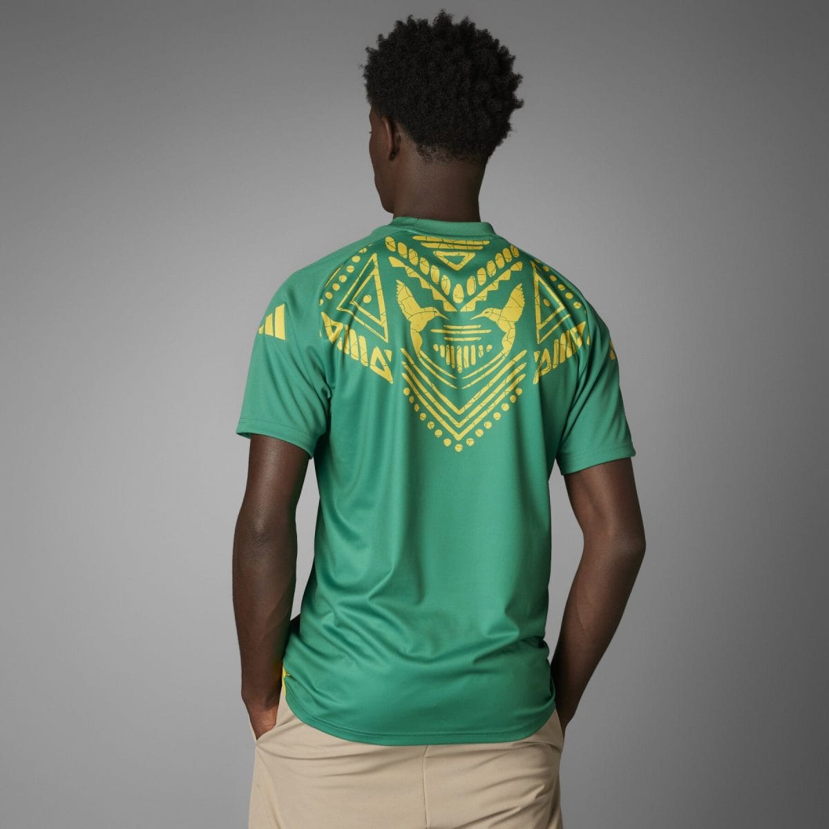 Person wearing a green shirt with a large yellow geometric design on the back, standing with beige shorts, seen from behind.