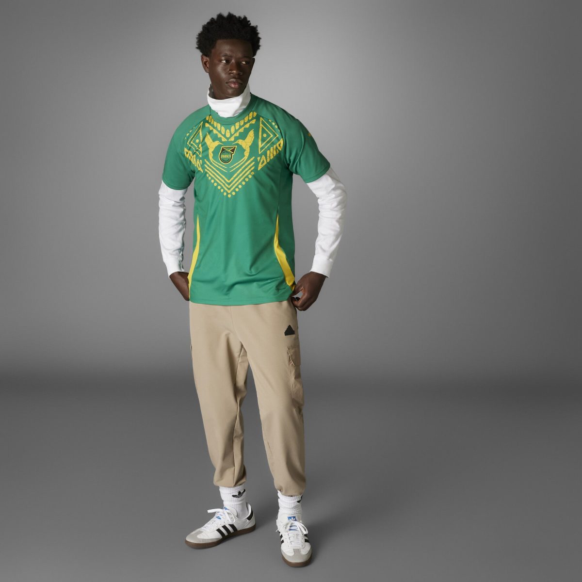 Full-body portrait of a man in a green geometric-patterned T‑shirt over a white long-sleeve top, beige pants, and white sneakers against a gray background.