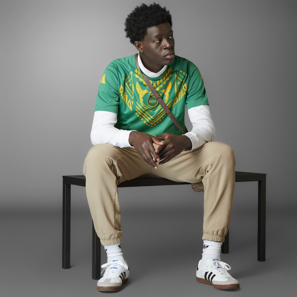 Young man sitting on a black bench, wearing a green graphic T-shirt over a white long-sleeve shirt and beige pants, looking to the side.