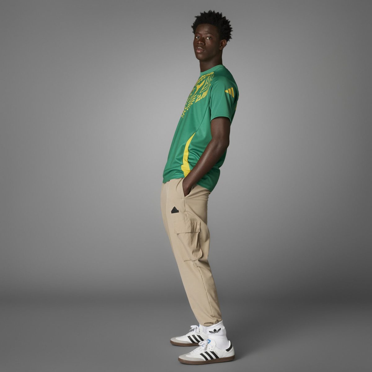 Side view of a man in a green graphic T‑shirt, beige cargo pants, and white sneakers against a gray studio backdrop.