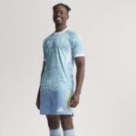 Smiling man in a light blue patterned soccer jersey and shorts, standing against a neutral background.