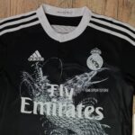 Black Real Madrid jersey with white collar and three white stripes on shoulders, featuring Adidas logo, Real Madrid crest, and 'Fly Emirates' sponsor with a dragon graphic in the center.
