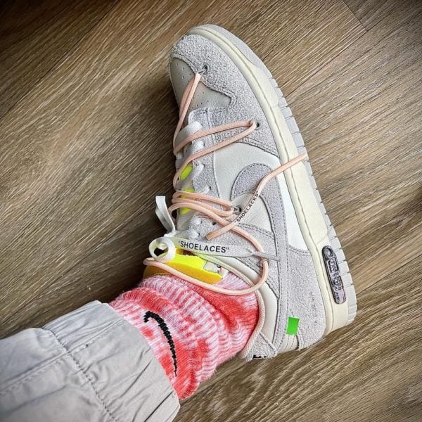 Close-up of a light gray athletic sneaker with peach-pink laces on a wooden floor, worn with a pink tie-dye sock lined with light gray pants.